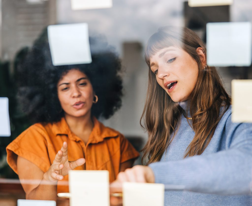 hiring local staff: females brainstorming in the office with post-its on w clear acrylic board.