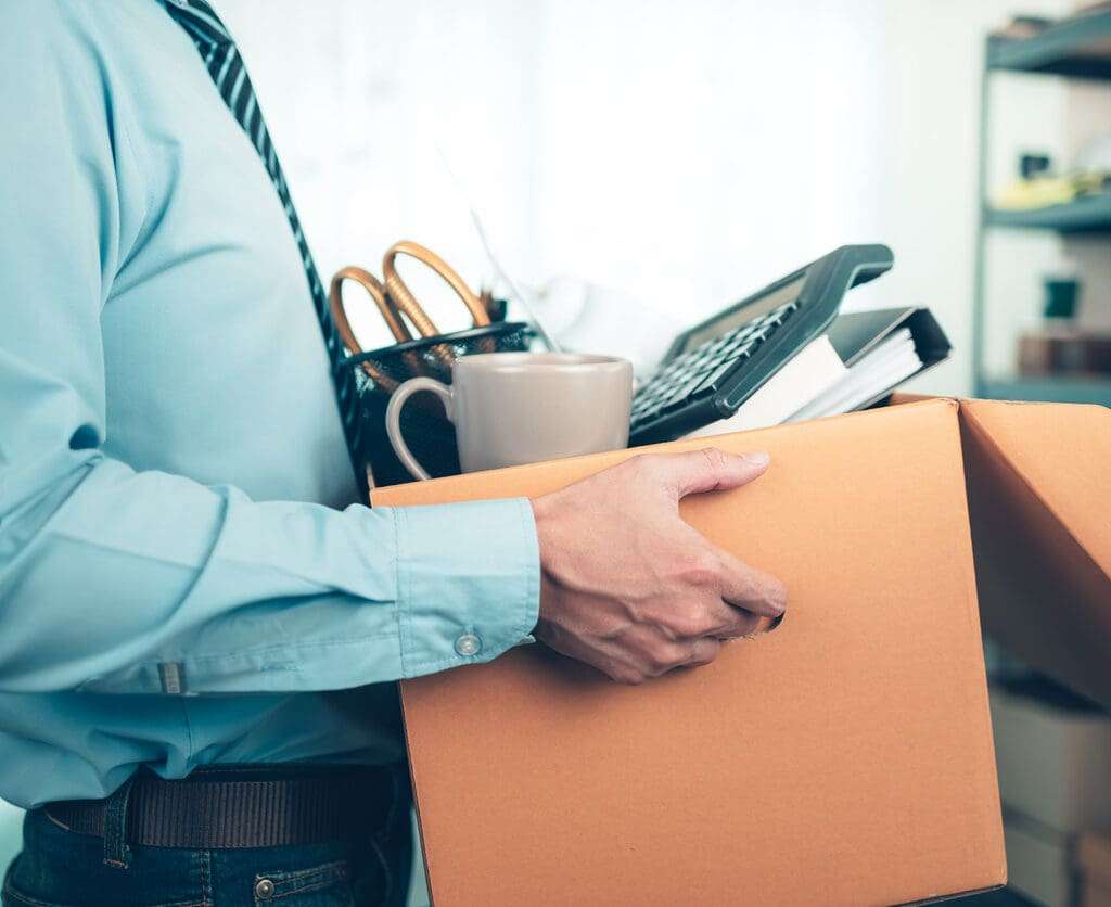 Man holding a cardboard box with, dossier, alarm clock, coffee cup, calculator and drawing tube.